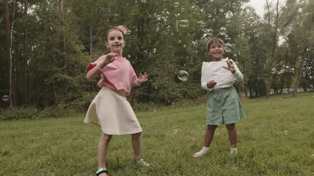 Low angle full of two multiethnic five-year-old girls smiling, dancing and catching soap bubbles in summer park on lawn