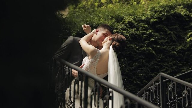 Newlyweds. Lovely young caucasian bride and groom stay on stairs in park making a kiss. Happy wedding couple. Man woman in love. Summer. Bride in gorgeous luxury wedding dress. Bridegroom in jacket
