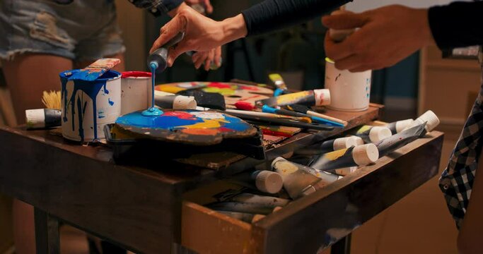 Artist's drawer, painter with scattered, disorganized paints in tube, brushes lying unwashed, artist's mess in paint store, studio, stuffed cabinet, woman squeezes color onto wooden palette