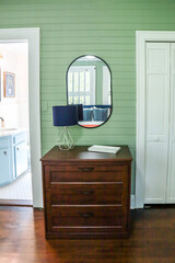 A simple dark wood stained dresser in a guest bedroom with green panel shiplap walls and a black rimmed mirror and small lamp