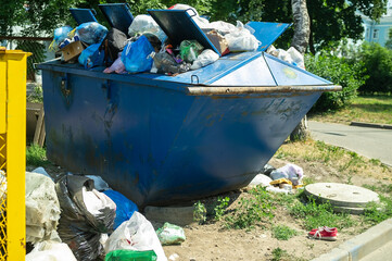 Bags of unsorted waste in and around an overflowing metal container in a city. Incorrect disposal...
