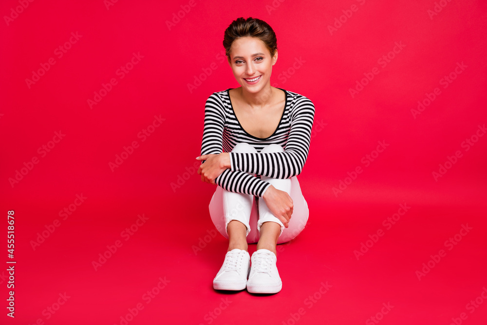 Poster Photo of pretty charming young lady wear striped clothes smiling sitting floor embracing knees isolated red color background