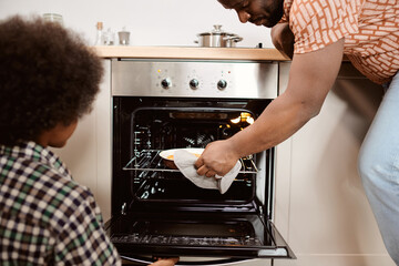 Young African man taking hot baked apple pie out of electric oven in front of his little son - Powered by Adobe