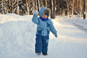 Fototapeta na wymiar A kid in warm winter clothes walks through a snow-covered park and throws a snowball
