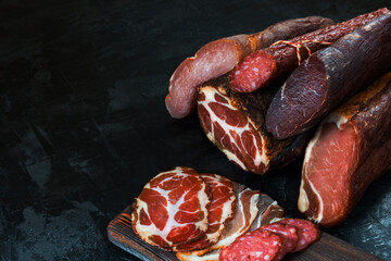 Whole and sliced meat of ham, sausage, prosciutto, jamon, bacon. Meat antipasto, banner, menu, black background. Copy space.