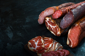 Whole and sliced meat of ham, sausage, prosciutto, jamon, bacon. Meat antipasto, banner, menu, black background. Copy space.