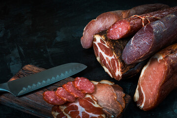 Whole and sliced meat of ham, sausage, prosciutto, jamon, bacon. Meat antipasto, banner, menu, black background. Copy space.