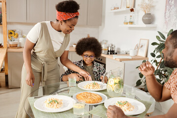 Young African woman putting plate with pasta on served table by her little son during family dinner