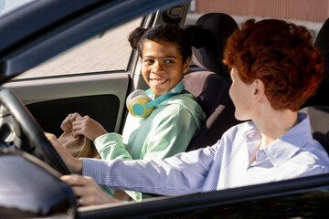 Happy mixed-race girl looking at young woman while both going to school by car