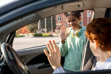 Smiling schoolgirl waving hand to her mom while getting out of car before going to school