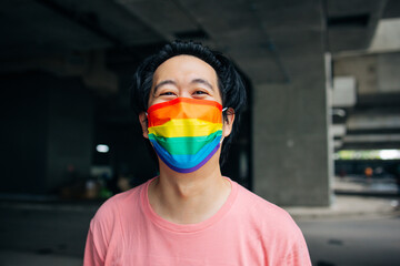 Young happy Asian man wearing a rainbow patterned mask representing LGBT support and pride during Covid-19 or Coronavirus - obrazy, fototapety, plakaty