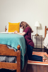 A young girl kneeling and praying to God at the side of a bed in her bedroom