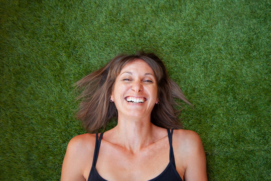 Mujer sonriendo echada sobre su espalda en el cesped verde. Mujer de pelo marr&oacute;n y camiseta de tirantes negro. Retrato de chica