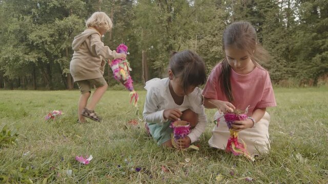 Wide shot of diverse gradeschoolers picking up candy fallen out of broken pinata in green park