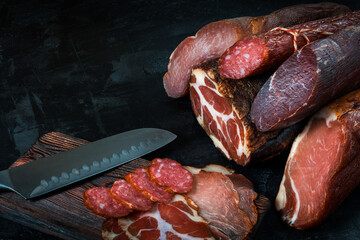 Whole and sliced meat of ham, sausage, prosciutto, jamon, bacon. Meat antipasto, banner, menu, black background. Copy space.