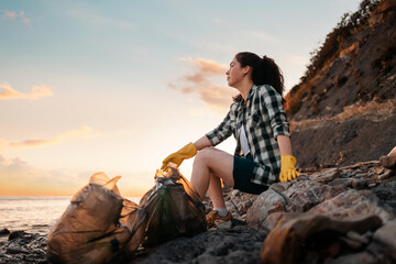 World environment day. A female volunteer sits on the beach with bags full of garbage. In the background, the sea and the sunset. Copy space. Bottom view. The concept of cleaning the coastal zone