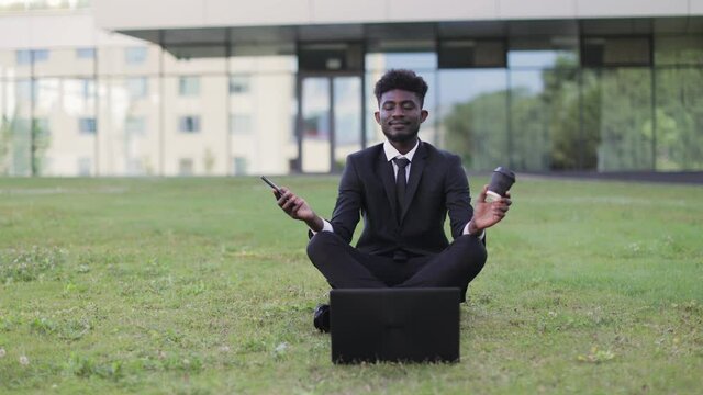 Office work and mindfullness. Handsome 30-aged African young business man meditating outside after work in front of company building, sitting on the grass with phone and coffee cup, with eyes closed