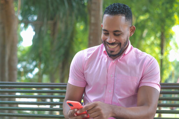 Naklejka na ściany i meble attractive dark-haired man of Dominican origin sitting in the park writing a message on his phone and laughing gesture, wearing a pink shirt