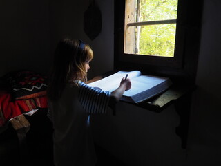 A girl of 8 years old with long blond hair near the window writes into a book. A room with...