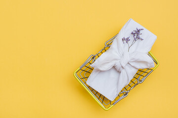 Eco-friendly furoshiki gift and dried flowers in a metal shopping basket on a yellow background. Black Friday, gift sales. Top view. Copy space