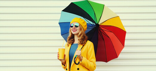 Autumn portrait of happy cheerful smiling young woman with colorful umbrella and cup of juice wearing a yellow coat and beret on a white background