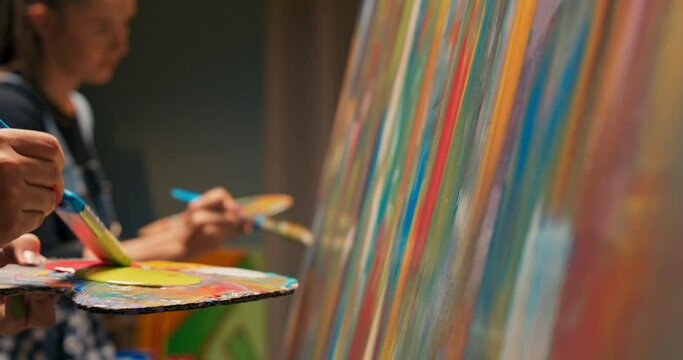 Painting together, creating a picture on a canvas located on an easel, women take red and yellow paint from a wooden palette and boldly paint two-colored stripes with a brush
