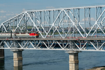 An electric train travels across a railway bridge across a river in an industrial city.