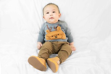 Fototapeta premium Cute 4 months little asian baby boy in stylish outfit and baby boots sitting on white blanket cool and happily at home,kid fashion.