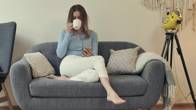 Young adult female person sits on cozy sofa at her apartment with smartphone and coffee cup in hands. Bare feet woman scrolls and browses, sips drink. Charming girl texting while lying on comfy couch.