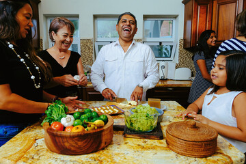 Cheerful mature man laughing and cutting cheese while cooking meal with family in kitchen at home