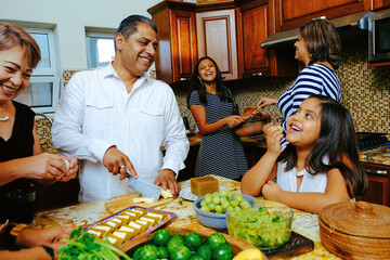Smiling girl eating grape while looking at father cutting cheese around family preparing appetizer together in kitchen during weekend