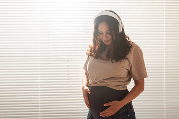 Happy pregnant woman with curly hair listening music in headphones, copy space.