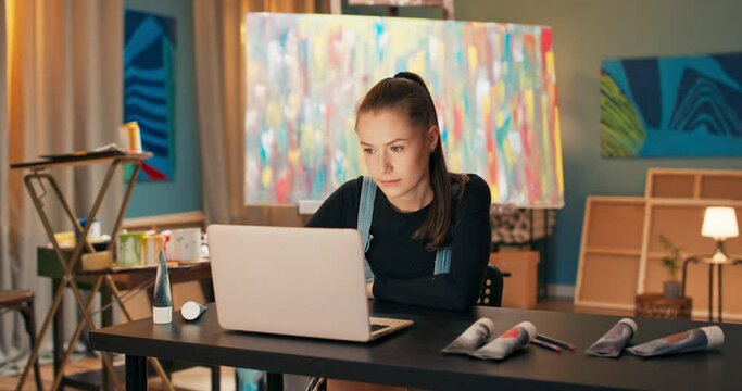 A smiling brunette sits at a desk in front of a laptop in art studio and displays paintings for sale, around lies a brush, watercolor paints in a tube, in the background a multi-colored painting