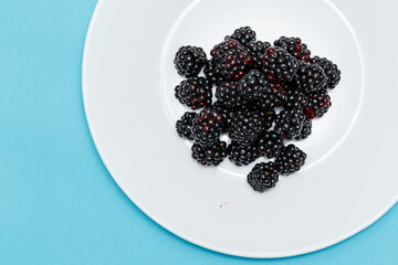 A white plate with blackberries on a blue background. Useful forest fruits. Super food. Concept of healthy eating. top view. Close-up. Macro. Blackberry berries with water drops. Flat lay