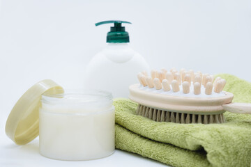 Double-sided massage brush for body brushing lies on green bamboo towel on white background. Dispenser cream and open jar of anti-cellulite body scrub. Materials for spa treatments