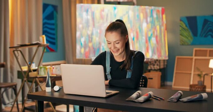 Artist, dressed in work clothes, sitting in studio, behind painting on canvas standing on easel, girl has laptop in front of her, she is talking through social media via webcam with manager