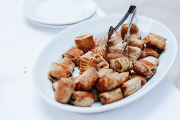 Close-up appetizers with puff pastry and sausages on porcelain plate on white setting table. Buffet during a gathering in a restaurant.