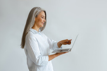 Smiling grey haired mature Asian businesswoman in white blouse points onto screen of modern laptop...