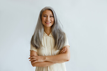 Portrait of smiling Asian woman with long hoary hair and crossed arms standing on light grey...