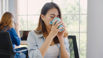 Cheerful asian young businesswoman sitting in office drinking coffee. Attractive asian creative woman holding a cup of coffee while working in workplace.