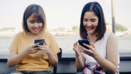 Fototapeta premium Young Asian women close friend tourist with casual enjoy with smartphone share photo in online social media in front of view port river at cafe city town, Lifestyle tourist travel holiday concept.