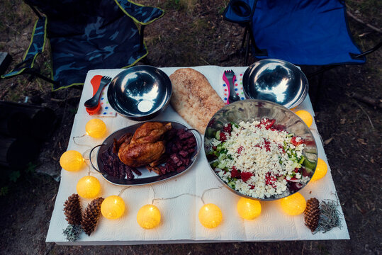 Two chairs and a camping table with dinner for two - salad, roast chicken, bread and decoration of cones and lights and lichen at dusk