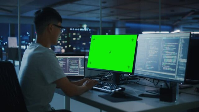Night Office: Young Japanese Man in Working on Green Screen Chroma Key Desktop Computer. Programmer Typing Code, Creating Modern Software, e-Commerce App Design. Over Shoulder