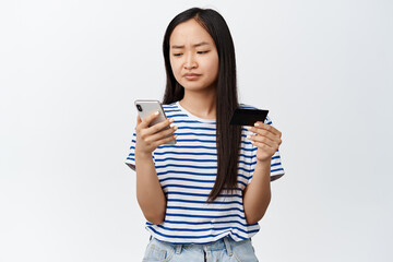 Fototapeta premium Asian girl has difficulties with online shopping app, problem during payment, frowning at mobile phone screen, holding credit card, standing over white background