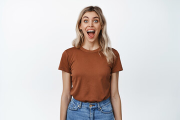 Fototapeta premium Image of blond girl looking surprised and excited, open mouth and widen eyes impressed, standing in t-shirt and jeans over white background
