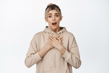 Image of emotional young woman, gasping affectionate, feeling touched, holding hands on heart, fond of something, standing against white background