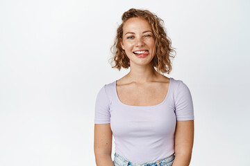 Positive blond girl shows tongue and winks at camera, makes happy and funny face, stands against...
