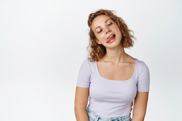 Funny blond girl showing tongue, fooling around and squinting, standing against white background