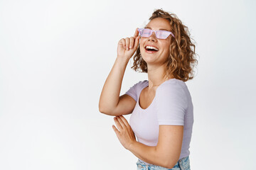 Stylish blond girl laughs in her sunglasses, smiles and looks happy, stands against white background