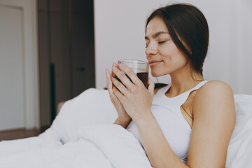 Side view young woman in white tank top sit in bed hold drink smell coffee from transparent glass rest relax spend time in bedroom lounge home own room house wake up dream be lost in reverie good day.